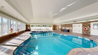Located at 6142 Merlot Dr in Highland, NY, this single-floor amenity facility offers a comprehensive suite of wellness and recreational spaces spanning a generously sized footprint. The entrance greets visitors with a bright, airy hallway finished in large-format tile flooring and accented by an elegant brass orb pendant light, setting a polished tone throughout.

The centerpiece of the facility is a spacious indoor pool enclosure featuring a freeform pool with an adjacent hot tub. Floor-to-ceiling windows line the perimeter, flooding the space with natural light and offering views of the outdoor patio area beyond. Stone accent walls and a vaulted beadboard ceiling lend the natatorium a refined, resort-like ambiance.

The exercise room is thoroughly equipped with a wide array of Life Fitness cardiovascular machines, including treadmills and ellipticals, as well as strength training equipment and cable machines. Natural light enters through multiple windows, and wall-mounted televisions provide entertainment. Adjacent to the main gym is a dedicated studio room with hardwood-style flooring, full-length mirrored walls, and interior windows overlooking the fitness floor — ideal for yoga, stretching, or group classes.

A comfortable lounge area with tile flooring, upholstered seating, decorative artwork, and a wall-mounted TV provides a welcoming space to relax, while connecting hallways ensure smooth circulation throughout the facility.
