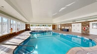 Located at 6142 Merlot Dr in Highland, NY, this single-floor amenity facility offers a comprehensive suite of wellness and recreational spaces spanning a generously sized footprint. The entrance greets visitors with a bright, airy hallway finished in large-format tile flooring and accented by an elegant brass orb pendant light, setting a polished tone throughout.

The centerpiece of the facility is a spacious indoor pool enclosure featuring a freeform pool with an adjacent hot tub. Floor-to-ceiling windows line the perimeter, flooding the space with natural light and offering views of the outdoor patio area beyond. Stone accent walls and a vaulted beadboard ceiling lend the natatorium a refined, resort-like ambiance.

The exercise room is thoroughly equipped with a wide array of Life Fitness cardiovascular machines, including treadmills and ellipticals, as well as strength training equipment and cable machines. Natural light enters through multiple windows, and wall-mounted televisions provide entertainment. Adjacent to the main gym is a dedicated studio room with hardwood-style flooring, full-length mirrored walls, and interior windows overlooking the fitness floor — ideal for yoga, stretching, or group classes.

A comfortable lounge area with tile flooring, upholstered seating, decorative artwork, and a wall-mounted TV provides a welcoming space to relax, while connecting hallways ensure smooth circulation throughout the facility.