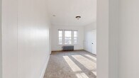 This single-floor unit offers a clean, functional layout with a bright and neutral aesthetic throughout. The living spaces are finished with plush wall-to-wall carpeting and crisp white walls, creating a light and airy atmosphere. Multiple rooms benefit from natural light streaming through gridded double-hung windows, with classic cast-iron radiators positioned beneath several of them, highlighting the unit's period character.

The carpeted bedrooms are generously proportioned, with one featuring a trio of windows that cast broad bands of sunlight across the floor. Closet storage is well-considered, with dedicated closets accessible through paneled white doors. One bedroom includes a double-door configuration offering access to both a closet and an adjoining hallway.

The kitchen is thoughtfully updated, featuring white shaker-style cabinetry, a subway tile backsplash, dark granite-look countertops, a matte black faucet, and light wood-look flooring. A gas range and stainless refrigerator complete the space, alongside a charming glass-panel built-in cabinet that adds vintage character.

The bathroom retains its classic appeal with white subway tile walls accented by bold dark border tiles, a full soaking tub, and a bright window providing natural ventilation and light over wood-look tile flooring.