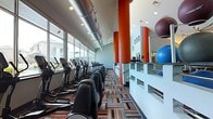 This contemporary fitness center is designed to inspire and motivate with a dynamic, energetic aesthetic. The main cardio floor is expansive, lined with a generous row of treadmills, ellipticals, and stationary bikes positioned along a full wall of floor-to-ceiling windows, offering views to the outdoors and flooding the space with natural light.

Striking orange cylindrical columns punctuate the floor plan, adding bold visual contrast against the light blue and white walls. Exposed silver ductwork runs along the ceiling, lending the space an industrial-modern character that complements the vibrant, multi-colored geometric carpet underfoot — featuring a mix of orange, blue, and neutral tones that energize the environment.

A raised platform area along the interior side of the gym provides dedicated space for functional training and flexibility work, outfitted with stability balls and additional equipment. Recessed and track lighting supplements the natural light, ensuring the entire space is well-lit throughout the day. The open layout, high ceilings, and thoughtful zoning create a functional yet visually engaging fitness environment suited for a wide range of workout routines.