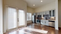 This well-appointed single-floor space offers a thoughtfully designed layout that combines style and functionality. The open-concept living and kitchen area is anchored by rich hardwood-style flooring that extends throughout, complementing the warm, neutral wall tones. Soaring ceilings enhance the sense of spaciousness, while large windows with horizontal blinds and a set of French doors flood the space with generous natural light, casting bright patterns across the floors.

The kitchen is a standout feature, outfitted with gray shaker-style cabinetry — including glass-front upper cabinets — paired with light stone countertops and a striking herringbone-pattern tile backsplash in muted metallic tones. A full suite of stainless steel appliances includes a gas range, microwave, refrigerator, and dishwasher, making the kitchen as practical as it is visually appealing. Under-cabinet lighting adds a refined finishing touch.

The bedroom area is generously sized, featuring ceiling fans and access to a private bathroom. The bathroom is appointed with a full soaking tub and shower combination, framed by floor-to-ceiling gray rectangular tile and accented with dark oil-rubbed bronze fixtures and built-in corner shelving. The overall design palette — neutral tones, dark hardware, and modern finishes — creates a cohesive, contemporary ambiance throughout the space.