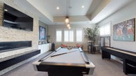 This single-floor entertainment space is designed for leisure and social gatherings, centered around a sleek, full-sized pool table beneath a striking cluster of industrial-style pendant lights. The tray ceiling adds architectural depth and a sense of elevated elegance to the room. A stacked stone accent wall serves as a dramatic focal point, framing a wall-mounted flat-screen television above a linear fireplace, lending warmth and texture to the space. Built-in white cabinetry below provides ample storage while maintaining a clean, polished aesthetic. Natural light filters generously through plantation-shuttered windows, brightening the neutral tones of the carpet and walls. A cozy lounge area with accent seating and coral throw pillows offers a relaxed retreat at one end of the room, while a high-top bar table and stools at the opposite side provide an ideal spot for casual dining or entertaining. A foosball table and additional gaming amenities further enhance the recreational atmosphere. Lush indoor greenery adds a refreshing organic touch. The overall ambiance strikes a refined balance between comfort and entertainment, making this a versatile and inviting space for both relaxed evenings and lively social occasions.