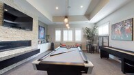 This single-floor entertainment space is designed for leisure and social gatherings, centered around a sleek, full-sized pool table beneath a striking cluster of industrial-style pendant lights. The tray ceiling adds architectural depth and a sense of elevated elegance to the room. A stacked stone accent wall serves as a dramatic focal point, framing a wall-mounted flat-screen television above a linear fireplace, lending warmth and texture to the space. Built-in white cabinetry below provides ample storage while maintaining a clean, polished aesthetic. Natural light filters generously through plantation-shuttered windows, brightening the neutral tones of the carpet and walls. A cozy lounge area with accent seating and coral throw pillows offers a relaxed retreat at one end of the room, while a high-top bar table and stools at the opposite side provide an ideal spot for casual dining or entertaining. A foosball table and additional gaming amenities further enhance the recreational atmosphere. Lush indoor greenery adds a refreshing organic touch. The overall ambiance strikes a refined balance between comfort and entertainment, making this a versatile and inviting space for both relaxed evenings and lively social occasions.