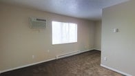 This single-floor residential space offers a straightforward and functional layout designed for comfortable everyday living. The main living area is finished with dark wall-to-wall carpeting that adds warmth underfoot, complemented by neutral beige walls that create a clean, understated backdrop throughout. A textured ceiling adds subtle visual interest to the room.

Natural light filters into the space through a large window fitted with horizontal blinds, offering views of the surrounding trees and greenery outside. Climate control is well-addressed, with a wall-mounted air conditioning unit positioned above the window for cooling, paired with a baseboard heating system running along the lower wall for year-round comfort.

The room's open, uncluttered layout provides flexible use of space, allowing for a variety of furniture arrangements to suit different needs. White baseboards frame the perimeter of the room, providing a clean finish between the walls and flooring. Electrical outlets are conveniently placed throughout the space.

Overall, this space presents a practical and livable environment with functional climate control features, natural light, and a calm, neutral aesthetic that serves as a versatile canvas for personalization.