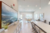 Located at 100 Edison Ave NW in Huntsville, Alabama, this single-floor community space offers a well-appointed layout encompassing a living room, kitchen, and office. The interior design embraces a clean, contemporary aesthetic characterized by light wood-look tile flooring, elevated ceilings, and a crisp neutral color palette throughout.

The expansive living room serves as the heart of the space, featuring recessed lighting, a wall-mounted flat-screen TV, and multiple seating arrangements including a striking live-edge communal table with modern chairs. A large landscape painting anchors one wall, adding a bold artistic focal point. Abundant windows along the perimeter flood the room with natural light and provide views of the surrounding outdoor areas, including a pool and neighboring buildings.

The kitchen is outfitted with dramatic dark cabinetry contrasted by white quartz countertops, a functional sink, and a large central island. Geometric pendant lights hang above, adding a refined modern touch, while ample counter space makes the area well-suited for gatherings.

The private office is a compact, functional room equipped with a wall-mounted display, a round table, and filing storage — ideal for focused work or small meetings.

French doors in the living area provide direct outdoor access, further enhancing connectivity to the broader property.