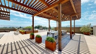 Located on Hatton Street in San Jose, California, this rooftop terrace offers a thoughtfully designed outdoor retreat with a range of amenities suited for both relaxation and social gatherings. The space is anchored by striking warm-toned wood pergolas strung with globe lights, providing partial shade over an al fresco dining area outfitted with long communal tables and chairs. The flooring transitions between large-format porcelain pavers and rich hardwood decking, creating distinct zones throughout the terrace. Generously sized lounge seating, featuring slatted wood-framed sectional sofas with deep charcoal cushions, offers a comfortable setting to take in panoramic views of the surrounding mountain ranges and San Jose skyline. Rust-patinated Corten steel planters filled with ornamental grasses are distributed throughout, adding natural texture and visual warmth to the space. Sun loungers positioned near the pergola provide additional relaxation options, while open sections of the terrace allow for flexible use. The perimeter railing frames unobstructed views of the verdant treeline and distant hills, completing the open-air ambiance. The overall design balances contemporary materials with natural wood elements, creating a refined and inviting outdoor environment.