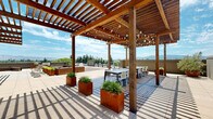 Located on Hatton Street in San Jose, California, this rooftop terrace offers a thoughtfully designed outdoor retreat with a range of amenities suited for both relaxation and social gatherings. The space is anchored by striking warm-toned wood pergolas strung with globe lights, providing partial shade over an al fresco dining area outfitted with long communal tables and chairs. The flooring transitions between large-format porcelain pavers and rich hardwood decking, creating distinct zones throughout the terrace. Generously sized lounge seating, featuring slatted wood-framed sectional sofas with deep charcoal cushions, offers a comfortable setting to take in panoramic views of the surrounding mountain ranges and San Jose skyline. Rust-patinated Corten steel planters filled with ornamental grasses are distributed throughout, adding natural texture and visual warmth to the space. Sun loungers positioned near the pergola provide additional relaxation options, while open sections of the terrace allow for flexible use. The perimeter railing frames unobstructed views of the verdant treeline and distant hills, completing the open-air ambiance. The overall design balances contemporary materials with natural wood elements, creating a refined and inviting outdoor environment.
