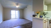 This single-floor residence offers a welcoming and well-lit interior designed for comfortable everyday living. The open living area is a standout feature, boasting vaulted ceilings that lend a sense of spaciousness and airiness to the room. A ceiling fan with a light fixture is centrally mounted, providing both style and function. Large sliding glass doors flood the space with natural light and open directly onto a private balcony, offering pleasant outdoor views and easy indoor-outdoor access. The flooring transitions from rich, dark hardwood near the entryway and kitchen area to plush wall-to-wall carpeting in the main living space, creating a warm and inviting atmosphere. A half-wall partition elegantly separates the kitchen from the living area, with a counter surface that serves as a convenient pass-through or casual dining spot. Neutral wall tones throughout complement the natural light and contribute to a clean, cohesive aesthetic. The overall ambiance is bright, open, and move-in ready, making this a well-appointed single-level space suited for a variety of lifestyles.