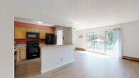 This single-floor unit offers a functional and straightforward layout designed for comfortable everyday living. The open-concept main area connects the kitchen and living room, creating a cohesive and practical space. The kitchen is well-equipped with light wood cabinetry, black appliances including a range, microwave, refrigerator, and dishwasher, along with laminate countertops and a double sink. A bold terracotta accent wall adds a touch of color contrast against the neutral cabinetry. A peninsula counter separates the kitchen from the living area, providing additional workspace and a casual dining option.

The living room features light laminate wood flooring, a textured ceiling, a wall-mounted air conditioning unit, and a baseboard heating system. A sliding glass door fills the space with natural light and provides direct access to an outdoor balcony with views of surrounding greenery.

The bedrooms are carpeted and quietly appointed, each featuring windows with views of mature trees and baseboard heaters. The bathroom is clean and functional, featuring a full tub-shower combination with a surround unit, wood-look flooring, and standard fixtures. Overall, the unit presents a practical and livable environment with a neutral palette throughout.