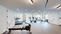 Located on Atlas Street in Baltimore, MD, this contemporary fitness center offers a well-appointed, single-floor layout designed to support a comprehensive workout experience. The space is characterized by a clean, modern aesthetic, with light-toned walls, durable rubberized flooring, and a distinctive slatted wood ceiling detail that adds warmth and architectural interest throughout.

The cardio zone is generously equipped with multiple treadmills, elliptical trainers, stationary bikes, and a rowing machine, all arranged to take full advantage of the expansive windows that line the perimeter. These large windows flood the space with natural light and provide views of the surrounding Baltimore streetscape, creating an energizing atmosphere for exercise.

A dedicated strength training area features a Life Fitness smith machine with bench, a full rack of dumbbells, and additional benches for free weight exercises. An accent wall showcasing abstract artwork adds a creative focal point to this zone, lending the space a polished, upscale feel.

An open studio area provides ample floor space for stretching, bodyweight training, or group fitness activities. A wall-mounted flat-screen display and warm wood-paneled walls enhance this multipurpose area. Recessed and linear LED lighting throughout ensures consistent, even illumination across all zones, completing this thoughtfully designed facility.