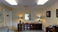 This single-floor space presents a well-appointed interior designed to make a strong first impression. The reception or display area serves as the focal point, featuring a broad, mid-century modern wooden credenza with open cubbies that showcase an array of trophies, awards, and accolades — a clear testament to the achievements associated with this space. Flanking a large wall-mounted mirror, a pair of sleek chrome-based table lamps cast a warm, inviting glow throughout the room, enhancing the professional yet approachable ambiance. The neutral-toned walls and carpeted flooring provide a clean, understated backdrop that keeps the focus on the displayed items. Overhead, flush-mounted light panels in a textured ceiling provide consistent illumination throughout the area. A window fitted with blinds allows for natural light control, contributing to the room's comfortable atmosphere. The overall design balances functionality with a polished aesthetic, making it well-suited for a professional office, real estate agency, or business headquarters where recognition and credibility are prominently on display.