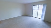 This single-floor space presents a clean, understated design with a welcoming and functional layout. The main living area is spacious and open, finished with light-toned wall-to-wall carpeting that adds warmth underfoot. Smooth, neutral-painted walls and a bright white ceiling create a calm, airy atmosphere throughout the room. A standout feature is the large sliding glass door, which floods the space with natural light and provides direct access to an outdoor balcony. The balcony overlooks a green, landscaped exterior, offering pleasant outdoor views. The sliding door is framed with a dark metal trim, providing a subtle contrast to the otherwise light interior palette. The room is free of visual clutter, making it highly adaptable for a variety of furniture arrangements and uses. Baseboard trim runs along the perimeter of the room, adding a clean finishing detail to the overall design. The combination of natural light, neutral finishes, and outdoor access gives this space a bright and inviting character that is both practical and comfortable.