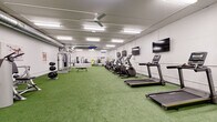 This single-level fitness center is designed to support a comprehensive range of workout routines in a clean, functional environment. The main workout area is expansive, featuring bright artificial turf flooring throughout, which provides a versatile surface suitable for cardio exercises, functional training, and agility drills. Overhead, rows of fluorescent lighting and ceiling fans ensure the space remains well-lit and ventilated throughout use.

The cardio zone is well-stocked with multiple treadmills, elliptical machines, a stair climber, and stationary bikes, with wall-mounted flat-screen televisions positioned for easy viewing during workouts. A dedicated strength training section houses a cable/multi-functional machine, weight benches, a dumbbell rack, and a power rack with weight plates. Exposed HVAC ductwork runs along the ceiling, contributing to the utilitarian aesthetic of the space.

Toward the rear of the facility, a mirrored wall spans a full section, visually expanding the space and supporting stretching, bodyweight exercises, or group training activities. Alongside the mirrors, a hardwood-style flooring area offers an alternative surface for floor-based workouts. Exercise balls, resistance accessories, and additional free weights are neatly organized nearby, completing this fully functional fitness facility.