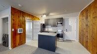 Located in Salt Lake City, Utah, this single-floor space blends updated modern finishes with characterful design details. The kitchen serves as a central highlight, showcasing dark gray shaker-style cabinetry with brushed gold hardware, a subway tile backsplash, and granite countertops that extend to a freestanding center island. A full suite of stainless steel appliances — including a refrigerator, range, microwave, and dishwasher — outfits the kitchen for everyday functionality. A gold-tone faucet adds a refined touch to the undermount sink. Adjacent to the kitchen, the living area features a striking knotty wood accent wall that spans nearly the full width of the room, providing warmth and texture against the neutral carpeted flooring and light-toned walls. Multiple windows dressed with horizontal blinds allow natural light to filter throughout. The bathroom is cleanly finished with full-surround white subway tile framing a soaking tub, complemented by brass hardware and a window for added ventilation and light. A laundry area with washer hookups is also present within the space. Overall, the unit offers a well-considered mix of contemporary updates and rustic character in a compact, efficiently laid-out single-level footprint.