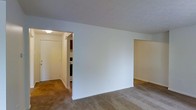 This single-floor space features a straightforward, functional layout with clean white walls and neutral-toned carpeting throughout the main living area. The open design allows for a natural flow between spaces, with a wide passageway leading into the kitchen, which is visible in the background and includes cabinetry and appliances. A separate doorway on the right side of the main room provides access to an additional area, offering a degree of spatial separation within the unit. The ceilings are smooth and painted white, contributing to a bright, airy feel, and a ceiling-mounted light fixture in the connecting hallway provides practical illumination. The overall aesthetic is simple and utilitarian, with neutral finishes that offer a blank canvas for customization. The carpeted flooring adds warmth underfoot, while the unadorned walls provide ample flexibility for furnishing and décor. The layout is efficient and well-connected, making it easy to navigate between the main living space, kitchen, and adjoining rooms.