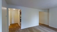 This single-floor space features a straightforward, functional layout with clean white walls and neutral-toned carpeting throughout the main living area. The open design allows for a natural flow between spaces, with a wide passageway leading into the kitchen, which is visible in the background and includes cabinetry and appliances. A separate doorway on the right side of the main room provides access to an additional area, offering a degree of spatial separation within the unit. The ceilings are smooth and painted white, contributing to a bright, airy feel, and a ceiling-mounted light fixture in the connecting hallway provides practical illumination. The overall aesthetic is simple and utilitarian, with neutral finishes that offer a blank canvas for customization. The carpeted flooring adds warmth underfoot, while the unadorned walls provide ample flexibility for furnishing and décor. The layout is efficient and well-connected, making it easy to navigate between the main living space, kitchen, and adjoining rooms.