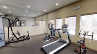 This fitness room offers a practical and functional space designed to support a variety of workout routines. The room is finished with neutral, warm-toned walls and dark carpet flooring, creating a focused environment suited for exercise. Recessed lighting throughout the ceiling provides even, consistent illumination across the space, while a row of windows along one wall allows natural light to filter in, enhancing the overall atmosphere.

A large wall mirror spans one side of the room, visually expanding the space and allowing users to monitor their form during workouts. The cardio equipment includes a treadmill and an elliptical machine, both positioned to take advantage of the natural light from the windows. A weight bench with an adjustable rack is also present, catering to strength training needs. Additional equipment visible in the mirrored reflection suggests further fitness options within the room.

A wall-mounted air conditioning unit ensures comfortable temperatures during use. The layout is thoughtfully arranged to maximize usable floor space, making the fitness room a convenient and well-appointed facility.