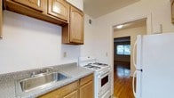 This single-floor residence offers a practical and well-organized layout designed for comfortable everyday living. The kitchen is equipped with warm-toned wooden cabinetry, both upper and lower, providing ample storage space. A speckled laminate countertop runs along the wall, complemented by a stainless steel single-basin sink and a white gas range with multiple burners and an oven below. A white tile backsplash lines the walls behind the counters, contributing to a clean and classic aesthetic. A ceiling vent ensures proper airflow throughout the space. The kitchen flows seamlessly through a doorway into an adjoining room featuring rich hardwood flooring that adds warmth and character. This connecting room benefits from natural light streaming in through a window, creating a bright and inviting atmosphere. The overall ambiance of the residence is straightforward and functional, with neutral wall tones and classic finishes that provide a versatile backdrop suitable for a variety of interior styles. The single-floor layout ensures easy accessibility and connectivity between spaces throughout the home.