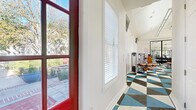 This fitness center offers a thoughtfully designed workout space spread across a single floor. The facility is accessed through a distinctive red entry door that opens to a welcoming corridor, where views of an outdoor patio and lush landscaping are visible through large, sunlit windows dressed with white plantation shutters.

The main workout area features vaulted ceilings with a ceiling fan and recessed lighting, lending an open and airy feel to the space. Crisp white walls and abundant natural light from multiple windows create a bright, energizing environment. A large wall mirror with black framing expands the visual depth of the room and reflects the full array of equipment within.

The floor is covered in a bold geometric-patterned tile in blue, white, and charcoal tones — a standout design element that adds a vibrant, modern character to the space. The equipment lineup is comprehensive, including treadmills, a rowing machine, a stationary bike, cable strength machines, resistance stations, free weights, and exercise balls neatly organized on a storage rack. Wall-mounted flat-screen televisions are positioned for visibility throughout the room.

The facility is well-suited for a full range of workouts, from cardio sessions to strength training, in a clean and visually dynamic setting.