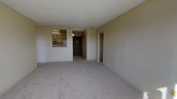 This single-floor residential unit offers a straightforward, functional layout with a clean and neutral aesthetic throughout. The main living area is spacious and open, featuring wall-to-wall carpeting in a soft, light beige tone that adds warmth underfoot. The walls are finished in a consistent neutral hue, contributing to a bright and cohesive atmosphere.

A pass-through window connects the living area to the kitchen, which is visible from the main room. The kitchen is equipped with standard cabinetry finished in a light wood tone, overhead lighting, a refrigerator, and counter space — providing a practical and efficient cooking environment.

From the living area, a hallway leads further into the unit, with multiple doors visible that suggest access to additional rooms such as a bedroom and bathroom. The entry door, finished in a warm wood tone, anchors the end of the hallway and provides a clear sense of depth and layout.

The space maintains a simple, no-frills design with standard ceiling heights and flush-mounted lighting. The overall ambiance is calm and utilitarian, well-suited for comfortable everyday living. The neutral palette and open arrangement of the main living zone make it an easily adaptable space for a variety of furnishing styles and personal tastes.