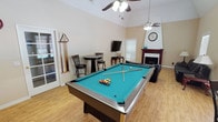 This single-floor recreational space is thoughtfully designed for leisure and social gatherings. The main area is anchored by a full-size pool table with a teal felt surface, complete with cue sticks and a rack, making it a natural centerpiece for entertainment. Light hardwood flooring runs throughout, complementing the warm, neutral-toned walls and creating a bright, welcoming atmosphere.

The lounge area features comfortable black leather sofas arranged around a glass-top coffee table, ideal for relaxing between games. A striking corner fireplace with a rich, dark wood mantle serves as a decorative focal point, topped with decorative accents and a wall clock. A wall-mounted flat-screen television is positioned nearby for added entertainment options.

Vaulted ceilings with ceiling fans enhance the sense of openness and help maintain comfortable airflow throughout the space. A bistro-style high-top table with bar stools provides a casual seating option, while French doors along one wall invite natural light and offer access to an outdoor area.

A compact kitchenette is also included, outfitted with white cabinetry, a sink, microwave, and full-size refrigerator — offering convenient refreshment access during gatherings. Windows with blinds allow for adjustable natural light throughout the space.