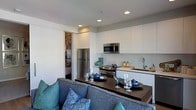 This single-floor space showcases a thoughtfully designed open-concept layout that seamlessly connects the living and kitchen areas. The kitchen is outfitted with sleek, floor-to-ceiling white cabinetry that maximizes storage while contributing to a clean, modern aesthetic. Stainless steel appliances, including a refrigerator, electric range, and built-in microwave, provide a polished, functional cooking space. A continuous dark-toned countertop extends from the kitchen into a dining or serving bar area, offering versatile surface space for both meal preparation and casual dining. The sink is positioned beneath a simple, modern faucet, and a dedicated section of the counter serves as a practical workspace. The living area flows naturally from the kitchen, anchored by a plush, grey sectional sofa accented with vibrant blue and teal throw pillows, adding a pop of color to the neutral palette. Recessed lighting throughout the ceiling ensures consistent, even illumination across the space. A sliding door visible in the background hints at additional rooms beyond, suggesting a practical use of space throughout the unit.