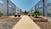 Located at 8116 E 151st St in Bixby, Oklahoma, this contemporary multi-building complex is defined by its thoughtfully designed outdoor common areas that serve as the centerpiece of the property. The exterior facades feature modern horizontal lap siding in neutral gray tones, with large, evenly spaced grid-style windows that allow abundant natural light throughout.

The expansive central courtyard runs the length of the complex, connecting multiple zones of outdoor living. String lights are strung overhead along the full corridor, creating an inviting atmosphere for evening gatherings. The courtyard is anchored by sleek steel pergolas that provide shaded dining and lounge areas, furnished with wicker and upholstered seating arranged for both intimate and group settings.

Raised concrete planters throughout the courtyard feature young trees and native plantings, adding greenery and visual interest to the hardscaped surfaces. Decorative gravel borders line the building perimeters, complementing the clean, low-maintenance landscaping aesthetic. Open lawn areas with well-maintained turf offer additional recreational space along the building edges.

Concrete walkways connect each outdoor zone seamlessly, providing clear and accessible circulation throughout the property. The overall design balances functionality with modern aesthetics, offering a variety of outdoor environments suited for relaxation, dining, and community engagement.