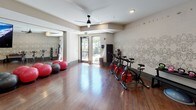 This single-floor fitness studio is thoughtfully designed to accommodate a variety of workout disciplines in a polished, welcoming environment. The space features rich hardwood flooring throughout, contributing warmth and a professional aesthetic that sets it apart from a typical gym setting.

The main exercise area is equipped with a row of spin bikes positioned along a decoratively wallpapered accent wall, which displays an intricate botanical or mandala-style mural paired with motivational text such as "Inhale, Exhale, Balance." Large floor-to-ceiling mirrors line one wall, visually expanding the room and supporting group fitness classes, yoga, and barre workouts. A ballet barre is conveniently positioned near the mirrors, and built-in cubby storage provides organized space for yoga mats and accessories.

Ceiling fans with a sleek, modern profile are installed throughout, promoting airflow during high-intensity sessions. Recessed lighting and a wall-mounted flat-screen TV add functional and entertainment value. Glass entry doors allow natural light to filter in from an adjacent outdoor area, offering glimpses of lush landscaping. The overall ambiance is clean, modern, and purpose-driven, balancing design sensibility with practical fitness functionality.