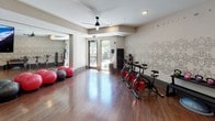This single-floor fitness studio is thoughtfully designed to accommodate a variety of workout disciplines in a polished, welcoming environment. The space features rich hardwood flooring throughout, contributing warmth and a professional aesthetic that sets it apart from a typical gym setting.

The main exercise area is equipped with a row of spin bikes positioned along a decoratively wallpapered accent wall, which displays an intricate botanical or mandala-style mural paired with motivational text such as "Inhale, Exhale, Balance." Large floor-to-ceiling mirrors line one wall, visually expanding the room and supporting group fitness classes, yoga, and barre workouts. A ballet barre is conveniently positioned near the mirrors, and built-in cubby storage provides organized space for yoga mats and accessories.

Ceiling fans with a sleek, modern profile are installed throughout, promoting airflow during high-intensity sessions. Recessed lighting and a wall-mounted flat-screen TV add functional and entertainment value. Glass entry doors allow natural light to filter in from an adjacent outdoor area, offering glimpses of lush landscaping. The overall ambiance is clean, modern, and purpose-driven, balancing design sensibility with practical fitness functionality.