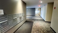 This single-floor interior hallway serves as a functional common area within the building, combining practical amenities with a clean, modern aesthetic. The corridor extends generously, providing ample circulation space for residents moving between units. Underfoot, a multi-toned patterned carpet in neutral hues of grey, beige, and brown adds visual texture while contributing to a quieter environment throughout the hallway.

A prominent built-in mailbox unit lines one wall near the corridor entrance, featuring numerous individual locked compartments organized in a grid arrangement — a centralized and efficient solution for mail distribution. The walls are finished in a soft neutral tone, complemented by accent walls in deeper grey, creating a calm and contemporary atmosphere.

Recessed ceiling lighting runs the length of the corridor, ensuring consistent illumination throughout. Emergency exit signage is clearly visible, reflecting attention to safety standards. The overall design is straightforward and practical, prioritizing clean lines and easy navigation, making this common area both welcoming and highly functional for everyday resident use.
