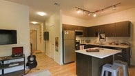 This well-appointed single-floor residence offers a thoughtfully designed layout that balances functionality with contemporary style. The open-concept kitchen serves as the heart of the space, showcasing rich dark cabinetry that extends to the ceiling, a sleek white countertop island ideal for casual dining or meal prep, and a full suite of stainless steel appliances including a refrigerator, microwave, and range. Directional track lighting illuminates the kitchen workspace, highlighting the clean lines and modern finishes throughout. The kitchen flows seamlessly into the living area, where warm hardwood flooring extends the length of the space, creating a cohesive and inviting atmosphere. The entry corridor, visible from the main living area, leads to additional rooms and storage, with built-in shelving providing practical organization. The living space is open and versatile, accommodating both relaxation and everyday activity. Light-toned walls throughout the unit reflect ambient light, keeping the interior feeling bright and airy. The overall design aesthetic is modern and urban, combining dark contrasting cabinetry with light surfaces and warm wood tones to create a balanced and welcoming environment.