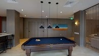 This single-floor recreational space is designed with both function and style in mind, centered around a full-size billiards table finished in rich dark wood with a deep navy felt surface. The table serves as the room's focal point, positioned beneath a trio of sleek black pendant lights that hang at varying heights, casting focused illumination across the playing surface while contributing a modern industrial aesthetic to the space.

The room features neutral-toned walls in warm grey and taupe hues, complemented by light-colored tile flooring that enhances the open, airy feel of the space. A horizontal abstract landscape painting adorns one wall, adding a vibrant pop of color and serving as a subtle artistic accent against the otherwise understated palette.

Recessed ceiling lights supplement the pendant fixtures, ensuring the room is well-lit throughout. Multiple doorways are visible along the back wall, suggesting the space connects to additional areas. Wall-mounted sconces provide ambient lighting along the perimeter. The overall ambiance is modern and inviting, with clean lines, thoughtful lighting design, and purposeful décor creating an atmosphere well-suited for leisure and social engagement.