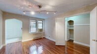 This efficiently designed single-floor unit offers a thoughtful layout encompassing a living room, kitchen, bathroom, and closet. The living room serves as the heart of the space, showcasing rich, polished hardwood floors that extend throughout. Neutral-toned walls, crown molding, and a curved archway add classic architectural character, while a track lighting fixture overhead provides adjustable illumination. A window unit air conditioner and a radiator ensure year-round comfort, and double windows allow natural light to filter in. An elegant arched doorway connects the living area to the walk-in closet, which features built-in shelving along multiple walls for organized storage, all set atop matching hardwood floors. The galley-style kitchen is accessed through a separate arched entry and is lined with warm wood cabinetry above and below on both sides, complemented by dark countertops and light tile flooring. It is equipped with a full suite of black appliances, including a refrigerator, stove, and dishwasher. The bathroom maintains a clean, classic aesthetic with white subway tile walls, a black-and-white mosaic tile floor, a pedestal-style vanity, a mirrored medicine cabinet, a soaking tub with shower, and a window for natural ventilation.