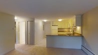 This single-floor space is designed with an open-concept layout that integrates the kitchen and living area into one cohesive environment. The kitchen is outfitted with white upper cabinetry providing ample storage, complemented by dark granite countertops that add contrast and a touch of elegance. A peninsula-style counter extends into the living area, creating a natural divide between the two spaces while maintaining visual openness. The kitchen includes a white refrigerator, a built-in microwave, and a stainless steel sink with a modern faucet. A tiled backsplash adds texture and detail to the cooking area. The flooring transitions from light ceramic tile in the kitchen to carpeting in the living area, delineating functional zones. Flush-mounted ceiling lights provide consistent, warm illumination throughout. The hallway area features a bi-fold closet door alongside a standard entry door, suggesting practical storage solutions near the entrance. Neutral wall tones and clean lines throughout contribute to a bright, understated aesthetic that is both functional and approachable.