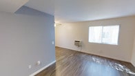This single-floor space offers a clean, functional layout designed for comfortable everyday living. The main living area is finished with rich, dark hardwood-style flooring that contrasts neatly with the crisp white baseboards and trim throughout. A distinctive slate-blue accent wall adds a touch of color and visual interest, while the remaining walls are painted in a neutral off-white tone, creating a bright and airy atmosphere. A ceiling fan is centrally positioned to support air circulation throughout the room. Climate control is provided by a wall-mounted HVAC unit, conveniently located beneath a large window fitted with vertical blinds. This window serves as the primary source of natural light, allowing sunlight to filter in and illuminate the space. The open floor plan creates a sense of spaciousness, with ample room to arrange furnishings according to personal preference. Multiple electrical outlets are distributed along the walls, ensuring practical usability throughout. The overall design is straightforward and well-maintained, making it a versatile and move-in-ready space suited for a variety of living arrangements.