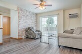 Located at 830 Union St in Platteville, Wisconsin, this generously sized single-floor unit offers six bedrooms and two full bathrooms across an expansive layout. The combined kitchen and living room serves as the heart of the space, featuring warm hardwood-style flooring, a ceiling fan with integrated lighting, and sliding glass doors that open onto a balcony overlooking a parking area framed by mature trees. The kitchen is equipped with rich cherry-toned cabinetry, a breakfast bar peninsula with seating, recessed lighting, and white appliances including a refrigerator and range. A second kitchen configuration also features matching cabinetry and ample counter space. The two bathrooms are well-appointed with dark-toned vanities, white countertops, mirrored medicine cabinets, and tub/shower combos, all finished with hardwood-style flooring and warm wood trim. The six bedrooms are carpeted and filled with natural light through window blinds, with several rooms featuring ceiling fans and baseboard heating. A central hallway connects the bedrooms, and two closets provide dedicated storage. The overall aesthetic is clean and functional, with neutral tones, consistent warm wood trim, and a practical floor plan well-suited for shared or multi-occupant living.