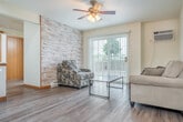 Located at 830 Union St in Platteville, Wisconsin, this generously sized single-floor unit offers six bedrooms and two full bathrooms across an expansive layout. The combined kitchen and living room serves as the heart of the space, featuring warm hardwood-style flooring, a ceiling fan with integrated lighting, and sliding glass doors that open onto a balcony overlooking a parking area framed by mature trees. The kitchen is equipped with rich cherry-toned cabinetry, a breakfast bar peninsula with seating, recessed lighting, and white appliances including a refrigerator and range. A second kitchen configuration also features matching cabinetry and ample counter space. The two bathrooms are well-appointed with dark-toned vanities, white countertops, mirrored medicine cabinets, and tub/shower combos, all finished with hardwood-style flooring and warm wood trim. The six bedrooms are carpeted and filled with natural light through window blinds, with several rooms featuring ceiling fans and baseboard heating. A central hallway connects the bedrooms, and two closets provide dedicated storage. The overall aesthetic is clean and functional, with neutral tones, consistent warm wood trim, and a practical floor plan well-suited for shared or multi-occupant living.