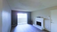 This single-floor space centers around a well-appointed living area that combines functional design with comfortable character. The room features hardwood-style flooring that runs throughout, providing a clean and cohesive foundation. A standout element is the white brick fireplace positioned along one wall, complete with a painted white mantle that serves as a natural focal point and adds a traditional, homey aesthetic to the space.

Natural light fills the room through a large sliding glass door dressed with vertical blinds, which leads to an outdoor balcony or patio area. The door is framed by a deep-toned accent wall that adds contrast and depth to the otherwise neutral color palette of soft whites and warm grays. The textured popcorn ceiling adds a layer of dimension overhead. The open layout of the living area allows for flexible furniture arrangement, making the space adaptable to various lifestyle needs. Overall, the room strikes a balance between classic architectural details and a practical, livable design.
