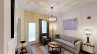 This single-floor space presents a comfortable and thoughtfully designed living area that balances modern aesthetics with functional furnishings. The room is anchored by a striking geometric chandelier with candelabra-style bulbs, serving as a focal point that draws the eye upward toward the smooth ceiling. Natural light enters through a glass-paneled door fitted with white blinds and is complemented by dark curtain panels that frame the windows, adding depth and contrast to the warm, light-toned walls.

The hardwood flooring runs throughout the living space, lending warmth and continuity to the room. A neutral-toned sofa pairs with a teal accent chair, creating an inviting seating arrangement centered around a round wooden coffee table set atop a patterned area rug. A tall floor lamp provides additional ambient lighting, while a large abstract wall artwork introduces a splash of color and artistic character to the space.

The overall ambiance is modern yet approachable, with a cohesive mix of industrial-inspired fixtures and soft furnishings that create a welcoming, lived-in atmosphere suitable for both relaxation and casual entertaining.