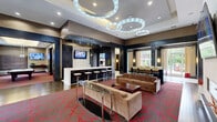 Park Lane at Garden State Park in Cherry Hill, New Jersey, presents a sophisticated community clubhouse designed with upscale amenities and a bold contemporary aesthetic. The space is anchored by a grand entry lobby featuring a striking reception desk with a textured stone facade, crisp white countertop, and illuminated Park Lane branding on a concrete-finish accent wall. Signature red-patterned carpeting flows throughout, tying together the various zones of the facility. The main lounge area is a showpiece, boasting dramatic circular chandelier installations overhead, a built-in bar with marble backsplash and bar seating, a wall-mounted flat-screen television, and a linear fireplace flanked by comfortable seating. A dedicated media room offers intimate theater-style seating with framed artwork and surround-sound cabinetry. The billiards room features a full-size pool table beneath sculptural pendant lighting, with large windows framing views of the surrounding greenery. A relaxed sitting area with hardwood flooring, curtained windows, and accent chairs provides a quieter retreat. Throughout the space, dark architectural trim, recessed lighting, and curated artwork contribute to a cohesive, upscale atmosphere designed for community gathering and recreation.