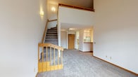 This two-floor residence offers a thoughtfully designed layout with warm wood accents and plush carpeting throughout. The main level centers around a spacious, open living area with vaulted ceilings that lend a sense of height and airiness. A wood-railed staircase leads to the upper loft level, which is illuminated by a skylight and features an open railing overlooking the living space below — ideal for use as a home office, reading nook, or additional lounge area. A ceiling fan adds comfort to this versatile upper space.

The galley-style kitchen is efficiently laid out with white cabinetry trimmed in warm wood tones, a full suite of white appliances including a refrigerator, range, and microwave, and a double sink beneath under-cabinet lighting. A pass-through counter connects the kitchen to the main living area, enhancing the open feel. In-unit laundry access is also present off the kitchen.

The living area benefits from a sliding glass door that opens to an outdoor balcony, filling the room with natural light and views of the surrounding greenery. A bedroom features a large window with a baseboard heater, while the bathroom includes a full tub/shower combination with tile flooring and a wood-trimmed vanity. Track lighting and recessed fixtures provide well-distributed illumination throughout the home.