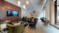 The Salon and Yard is a thoughtfully designed single-floor amenity space that blends industrial character with refined, contemporary furnishings. The open layout is anchored by soaring ceilings with exposed ductwork and structural elements, lending an airy, loft-like atmosphere throughout. Polished concrete floors run continuously underfoot, tying together the various seating zones with a cohesive, modern aesthetic.

The main lounge area features plush green velvet sofas and armchairs arranged around a glass-top coffee table, complemented by sculptural gold geometric pendant lights that add warmth and sophistication. A rich wood-paneled wall provides a dramatic backdrop for a mounted flat-screen display, while a vintage mail console lends a nod to the building's history.

Adjacent to the lounge, a dining area offers a round walnut table surrounded by gold velvet chairs, perfectly positioned beneath a curated gallery wall of black-and-white framed artwork in gold frames. A built-in wet bar with a beverage cooler and open shelving serves this area with ease.

Floor-to-ceiling black-framed windows and glass doors along one wall flood the interior with natural light and provide direct views and access to the outdoor yard, where additional seating is visible. The overall ambiance is social yet sophisticated, designed to encourage both relaxed gathering and casual conversation.