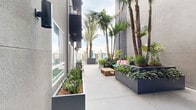 Located at 7650 Van Nuys Blvd in the San Fernando Valley neighborhood of Los Angeles, this outdoor amenity space delivers a resort-inspired atmosphere within a contemporary multi-story building. The grounds are organized into several distinct zones, each thoughtfully designed for relaxation and social gathering. A sparkling pool serves as the centerpiece, surrounded by a generous deck lined with orange lounge chairs and teal umbrellas, evoking a vibrant, California-casual aesthetic. Tall palm trees rise throughout the space, providing natural shade and reinforcing the Southern California character of the setting. Adjacent to the pool area, a sun deck offers additional seating through brightly cushioned chaise lounges, canopied swing beds, and bean bag chairs, all set against open sky views of the surrounding neighborhood. Intimate courtyard corridors wind between the building's facades, featuring raised planter beds filled with lush tropical and ornamental greenery. These walkways are furnished with teak wood benches and coffee tables adorned with bold, colorful cushions, creating cozy gathering spots. Oversized statement planters punctuate key intersections, adding sculptural interest. The building's exterior combines smooth gray and white stucco panels with large windows and metal-railed balconies, framing the outdoor spaces with a clean, modern architectural backdrop throughout.