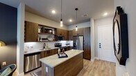 This thoughtfully designed single-floor residence blends contemporary aesthetics with functional living. The open-concept kitchen serves as a centerpiece, showcasing rich walnut-toned cabinetry with matte black hardware, a granite-topped island with pendant lighting overhead, a full suite of stainless steel appliances including a side-by-side refrigerator with ice and water dispenser, an electric range, built-in microwave, and dishwasher. A white tile backsplash and under-cabinet lighting add a clean, polished contrast to the warm wood cabinetry. Light wood-style flooring flows seamlessly throughout the space, lending continuity and warmth. The living area features a bold, dark accent wall that provides a dramatic backdrop for vibrant large-format artwork, complemented by a cognac leather sofa and stylish lighting. The bedroom continues the sophisticated palette with a deep navy accent wall, a ceiling fan, natural light from window panels with drapes, and direct access to the en-suite bathroom. The bathroom is finished with a granite-topped vanity, wood-toned cabinetry, a large mirror, and a tiled shower, maintaining the cohesive, upscale design language found throughout this well-appointed residence.