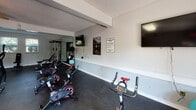This single-floor fitness center is designed to provide a comprehensive workout environment in a clean, functional layout. The space is accessed through a bright entryway featuring a set of white-framed French doors that flood the area with natural light and offer views of the surrounding greenery outside. A secondary glass-panel door provides an additional entry point, lending the space an open, welcoming feel.

The main exercise area is finished with durable dark rubberized flooring throughout, offering both safety and practicality for a range of workout activities. A large wall-to-wall mirror runs along one side of the room, visually expanding the space while allowing users to monitor their form during training sessions. The room is equipped with a dumbbell rack offering a variety of free weights, multiple adjustable workout benches, a cable machine, and an assortment of cardio equipment including stationary bikes and an elliptical-style machine. Wall-mounted flat-screen televisions are positioned throughout the space for entertainment during workouts.

Natural light filters in through double-hung windows fitted with blinds, complementing the overhead fluorescent lighting. The textured ceiling and clean, light-gray walls contribute to a no-frills, purpose-driven atmosphere. Posted exercise room rules and motivational wall art complete the functional aesthetic of this well-appointed fitness facility.