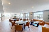Located on Eby Street South in Kitchener, Ontario, this well-appointed amenity lounge offers a generously sized, open-concept layout across a single floor. The space is designed with a contemporary aesthetic, featuring warm honey-toned hardwood-style flooring, soft grey walls, and recessed pot lighting throughout that creates a bright, welcoming atmosphere.

The main lounge area is thoughtfully furnished with multiple round glass and dark-topped tables surrounded by tan leather armchairs, offering flexible seating arrangements suited for both social gatherings and casual meetings. A comfortable leather sofa and loveseat grouping, anchored by a sleek dark coffee table, provides a relaxed lounge zone. A wall-mounted flat-screen television is positioned within one seating area, adding a functional entertainment element.

The fully equipped kitchen features white shaker-style cabinetry, white quartz countertops, stainless steel appliances including a refrigerator and over-the-range microwave, and a double-basin sink. Pendant lighting with modern dome fixtures illuminates the workspace, while a large window allows ample natural light to pour in.

Large windows and glass doors line the perimeter of the lounge, offering abundant natural light and direct access to an outdoor terrace with open city views. Curated artwork and indoor greenery add polished finishing touches throughout.