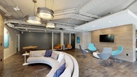 This single-floor communal space showcases a thoughtfully designed, open-concept layout that blends industrial and contemporary aesthetics. Exposed ductwork and plumbing runs along the ceiling, complemented by large perforated drum pendant lights that cast warm, inviting illumination throughout. Wood-look tile flooring grounds the space and flows seamlessly from zone to zone.

A striking curved sectional sofa serves as the centerpiece of the main lounge area, surrounded by an eclectic mix of seating options including modern swivel chairs in shades of blue and gray, and warm cognac leather chairs that add contrast. Decorative patterned partition screens subtly divide areas without closing off the open feel. A wood-paneled accent wall anchors a wall-mounted flat-screen TV and is flanked by vibrant aquatic-themed artwork that introduces pops of teal throughout.

A secondary lounge zone features paired dark sofas, striped area rugs, and wooden coffee tables with cylindrical ottomans, creating a more intimate gathering spot. The kitchenette area is equipped with a commercial-grade coffee and beverage station, microwave, and sink, offering practical amenities for occupants.

Floor-to-ceiling windows along one wall flood the space with natural light and provide views of the surrounding outdoor landscape, further enhancing the bright, airy ambiance of this versatile communal environment.