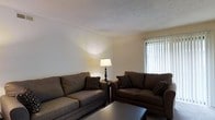 This single-floor space presents a welcoming living room as its focal point. The room is furnished with two generously sized sofas in warm brown tones, accented with patterned throw pillows that add a subtle decorative touch. A classic floor lamp with a cream shade anchors the seating area, casting a warm ambient glow across the neutral-colored walls. A low-profile coffee table sits at the center, completing the seating arrangement. Natural light filters into the room through a window dressed with vertical blinds, brightening the space and offering a connection to the outdoors. The textured ceiling adds a traditional architectural detail, while the overall layout of the room is practical and efficiently arranged. The color palette throughout is understated and cohesive, combining neutral walls with earth-toned furnishings to create a calm, inviting atmosphere. The space is thoughtfully arranged to maximize comfort and functionality, making the living area a relaxed and approachable environment suitable for everyday use.