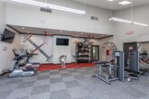 This fitness center at 6865 Bebout Dr in Norcross, Georgia, offers a comprehensive workout environment across a single floor. The space is divided into distinct functional zones, each designed to support a variety of exercise routines. Soaring vaulted ceilings in the main area create an open, airy atmosphere, complemented by a red ceiling fan and recessed lighting that ensure the space remains bright and well-ventilated.

The cardio zone features multiple TRUE-brand treadmills and an elliptical trainer, arranged along a wall lined with horizontal windows that invite natural light and outdoor views. Wall-mounted flat-screen televisions are positioned throughout the space, providing entertainment during workouts.

A dedicated strength training area houses a variety of selectorized weight machines and is enhanced by floor-to-ceiling mirrors that amplify the sense of space. An adjacent cycling zone features spin bikes on distinctive red flooring, set against a painted white brick accent wall adorned with a bold geometric mural, adding a stylish, industrial-chic character to the room.

A free weights section offers dumbbells and medicine balls stored on sturdy racks, rounding out the facility's comprehensive equipment offering. The overall flooring combines gray carpet tile with the striking red rubberized cycling area, creating clear visual distinctions between workout zones.