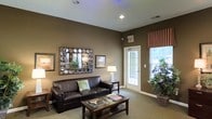 This single-floor space offers a thoughtfully designed interior that combines comfort and style in a cohesive, welcoming environment. The lounge area serves as a central gathering space, featuring rich earth-toned walls that create a warm and inviting ambiance. Dark leather seating is arranged around a wooden coffee table, providing a comfortable and functional setting for relaxation or conversation.

The room is adorned with carefully selected décor, including a decorative circular-framed mirror that adds visual interest to the wall, accompanied by framed artwork that enhances the overall aesthetic. A floor lamp and table lamp provide soft, layered lighting to complement the recessed ceiling lights above.

Natural light filters into the space through a glass-paneled exterior door and a window dressed with striped Roman shades, offering a pleasant connection to the outdoors. Lush potted plants are positioned near the entrance, introducing a fresh, natural element to the interior.

Neutral carpeting runs throughout, tying together the warm color palette and adding to the overall sense of comfort. The layout is open and well-organized, with clean white trim providing a crisp contrast against the darker wall tones. Overall, the space presents a polished, functional design suited for both everyday use and welcoming visitors.